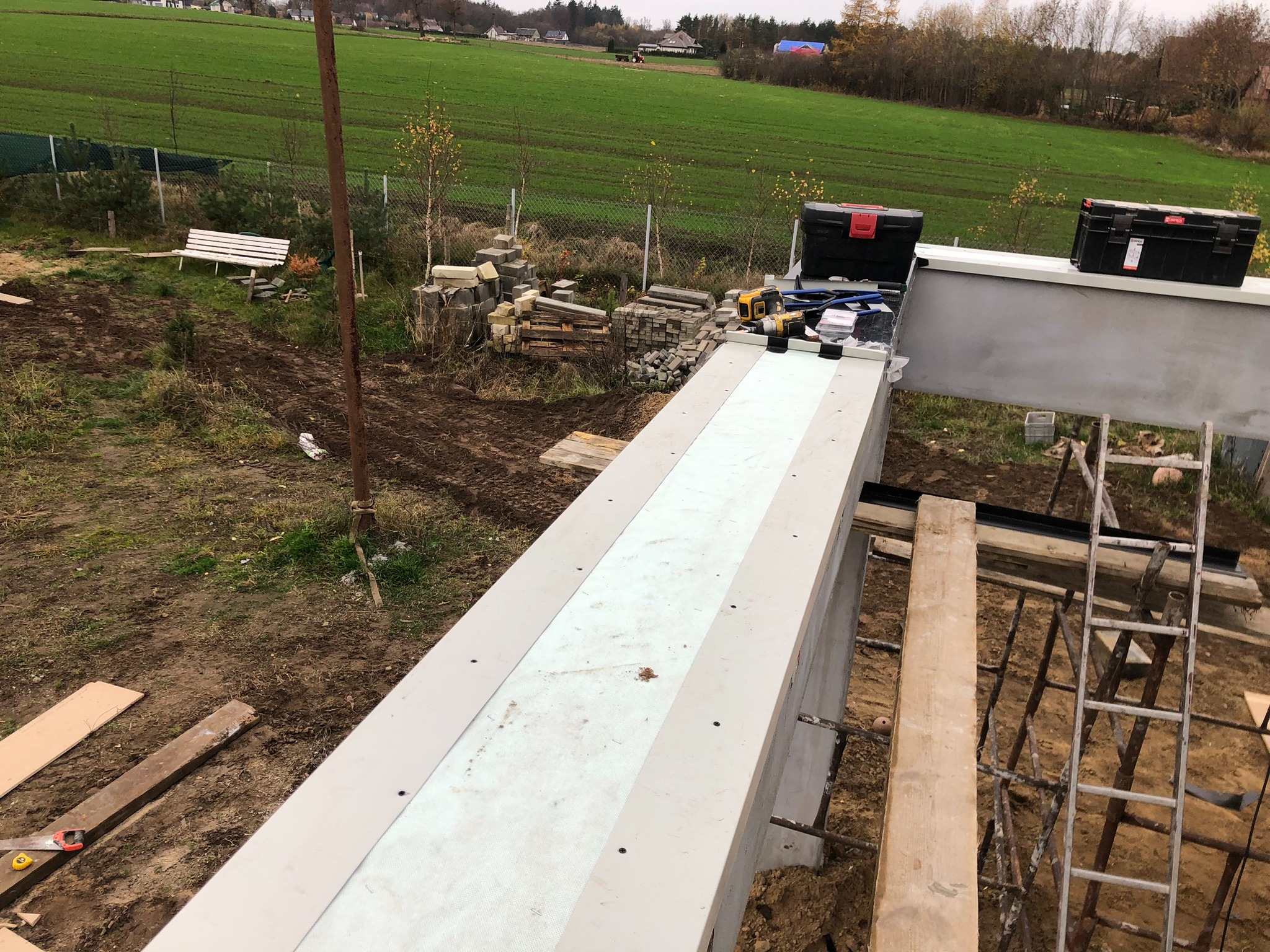 Widok z góry na konstrukcję betonową z izolacją termiczną w trakcie budowy, narzędzia i skrzynki narzędziowe na górze, drabina oparta o konstrukcję, w tle zielone pole i zabudowania.