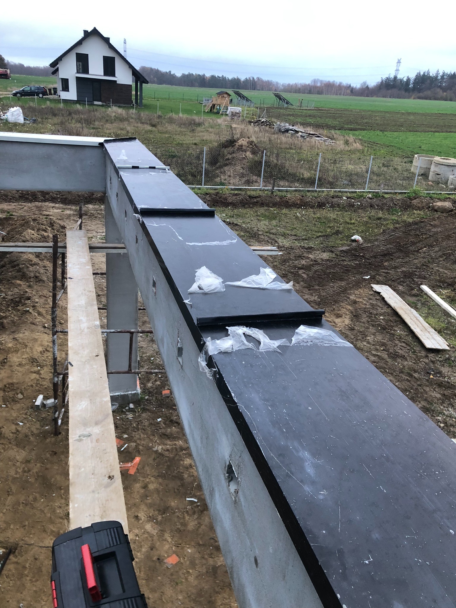 Betonowa konstrukcja dachu z czarnym wykończeniem, częściowo zabezpieczona folią, widok z góry, w tle dom jednorodzinny i krajobraz.