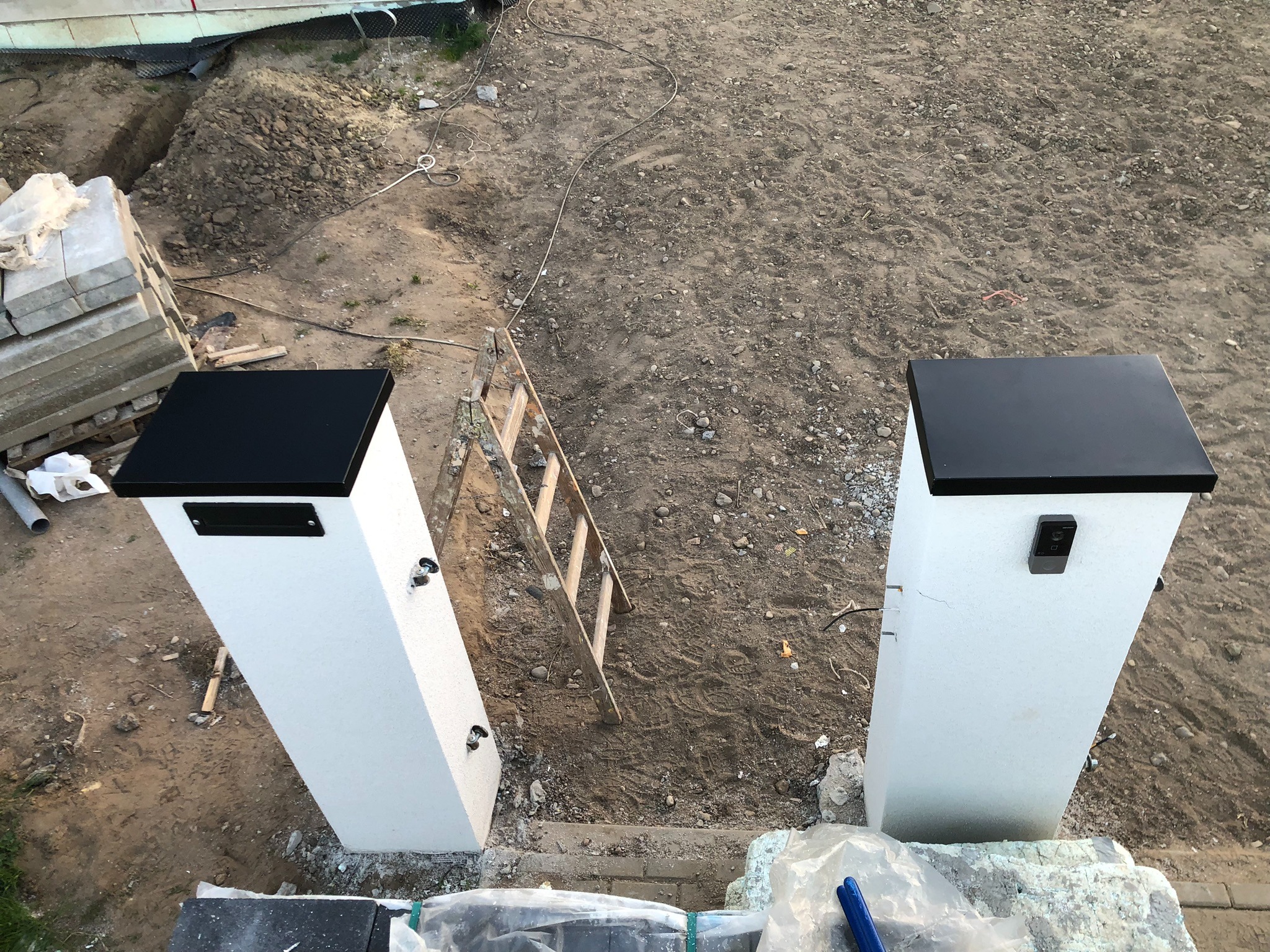 Dwa białe słupki ogrodzeniowe z czarnymi daszkami i domofonem, widok z góry na plac budowy z drabiną, ziemią i materiałami budowlanymi.