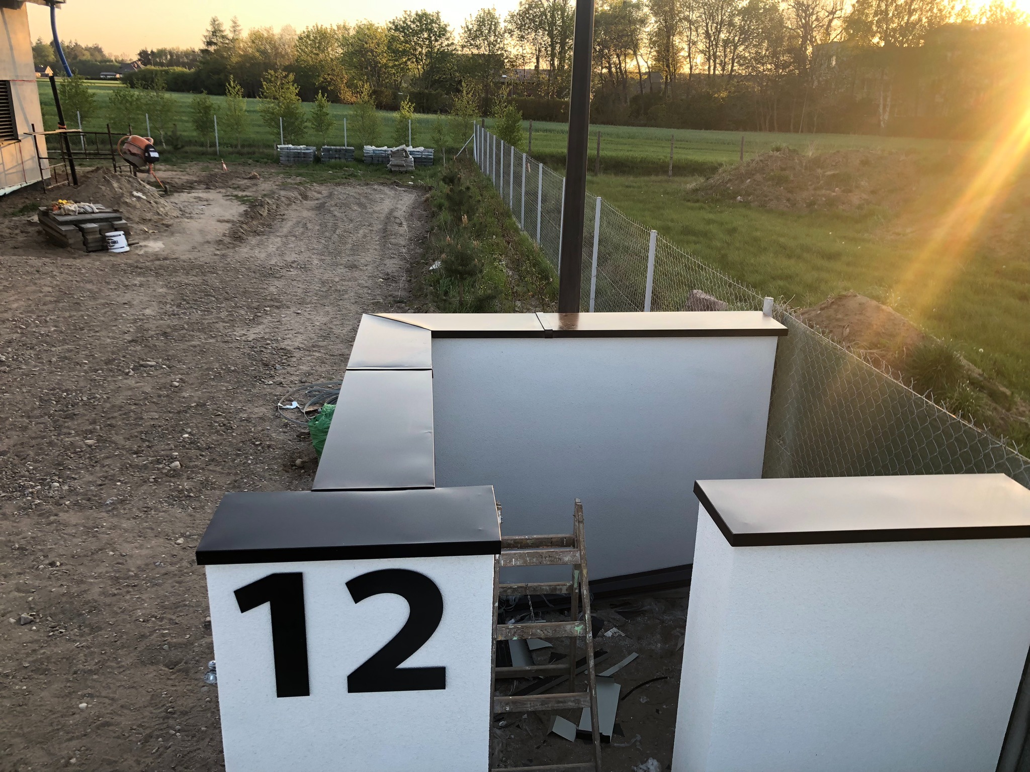 Wykończona elewacja domu z numerem 12, widoczna drabina i teren w trakcie prac wykończeniowych, w tle zieleń i zachodzące słońce.