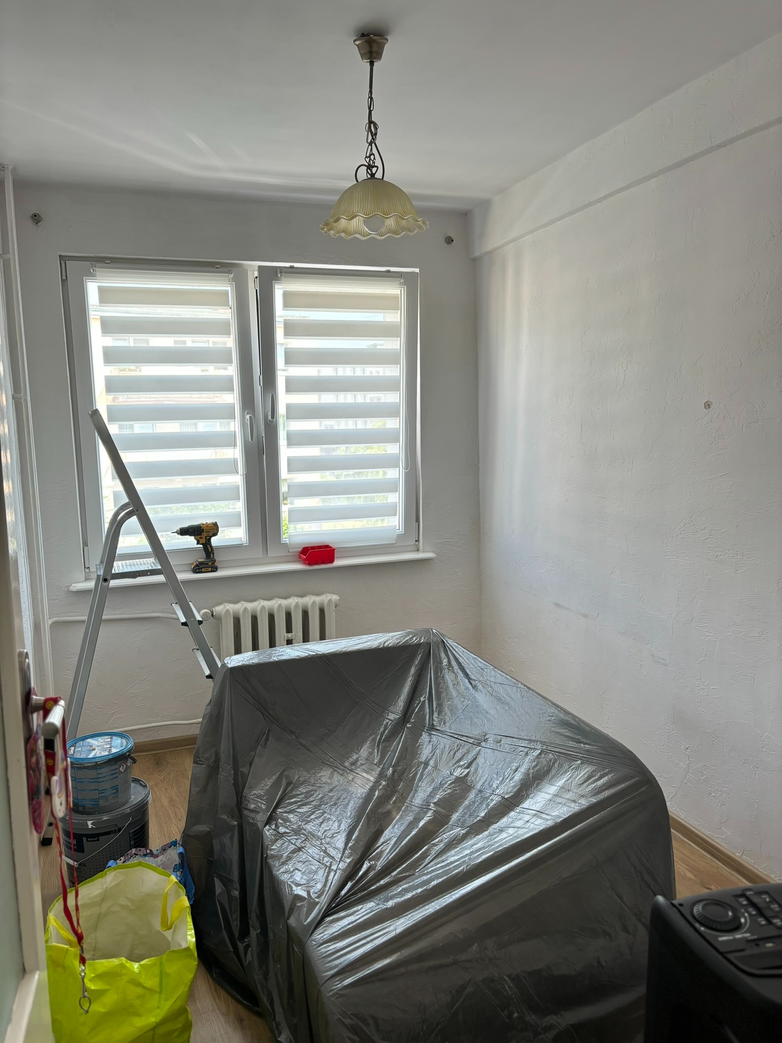 Pokój w trakcie przygotowań do malowania: meble przykryte folią ochronną, drabina oparta o okno z roletami, wiertarka na parapecie, puszki z farbą obok drzwi.