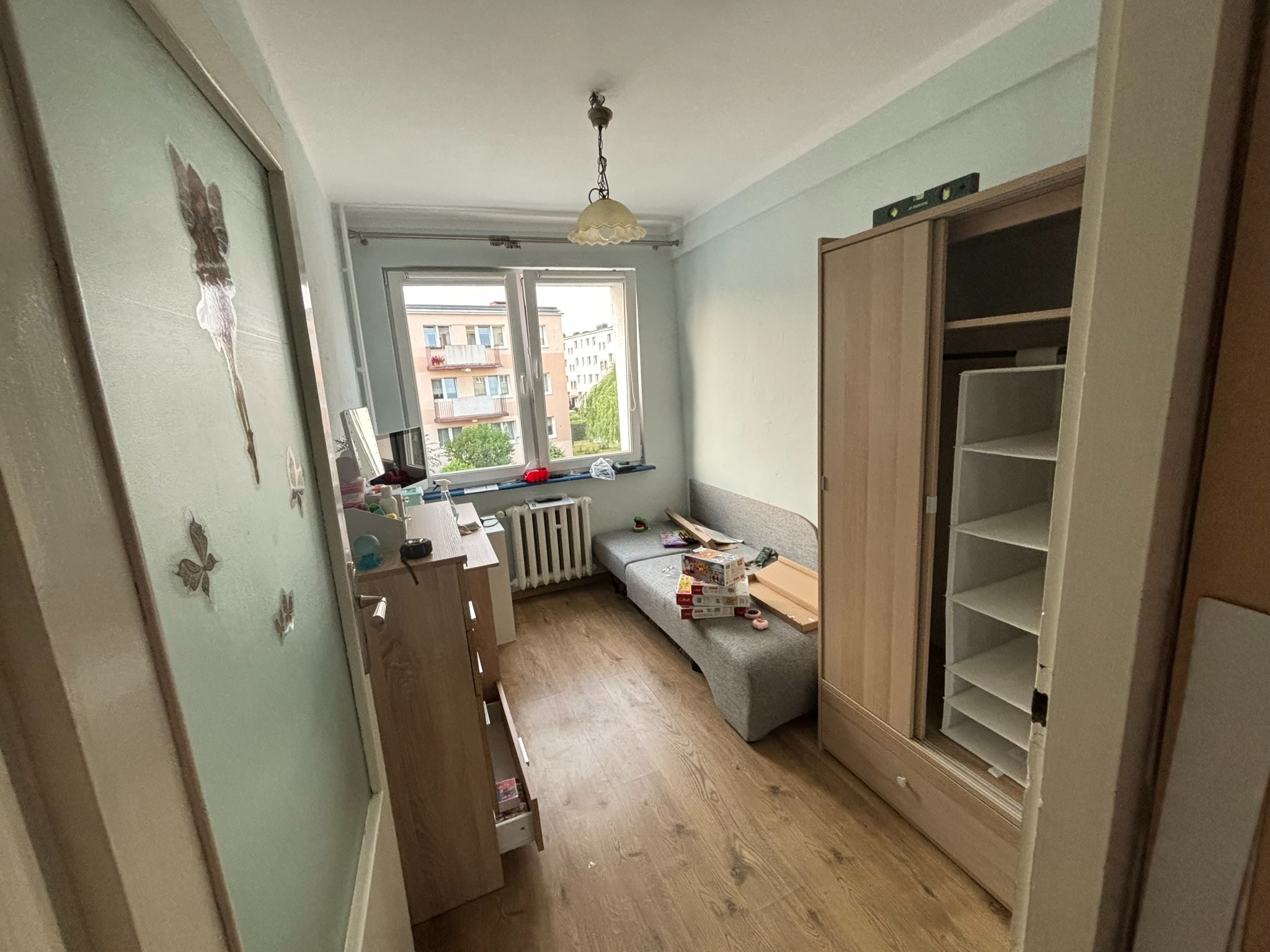 Pokój w trakcie drobnego remontu: widoczne meble, podłoga z paneli, okno z widokiem na blok i rozłożona sofa z pudełkami. Na szafie leży poziomica.