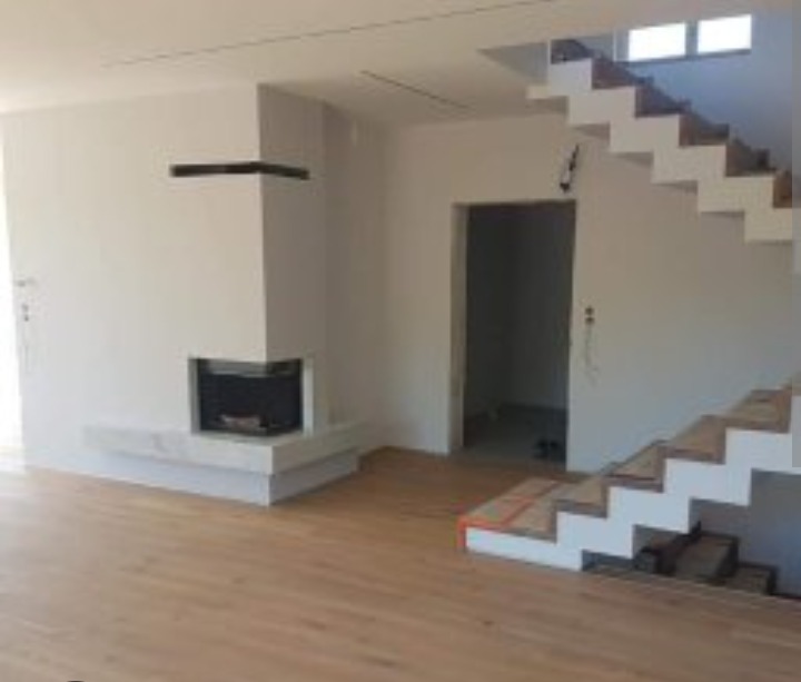 Nowoczesne wnętrze z drewnianą podłogą, białymi schodami i minimalistycznym kominkiem. Widoczne niedokończone prace wykończeniowe.