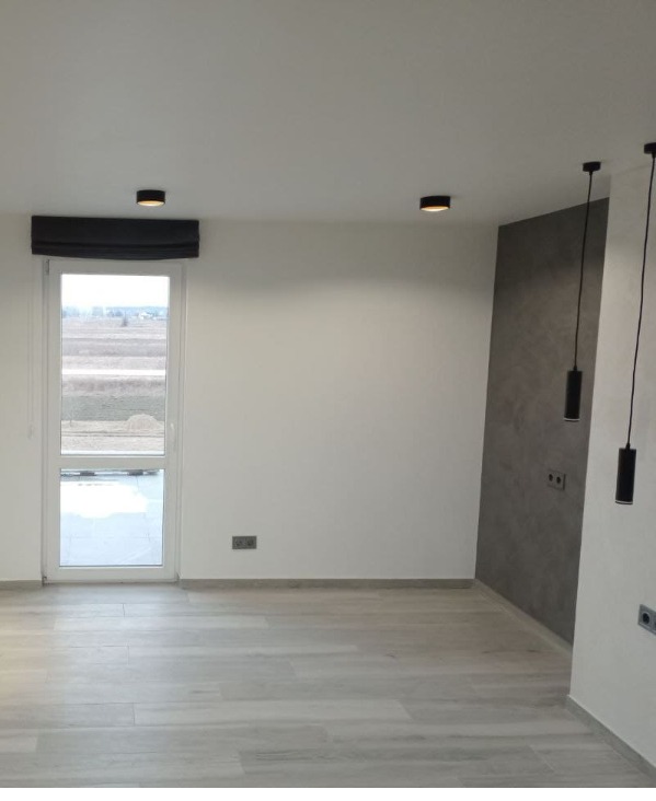 Minimalistyczne wnętrze: jasne panele, białe ściany, okno z roletą rzymską i nowoczesne lampy wiszące. Szara ściana akcentowa dodaje charakteru.