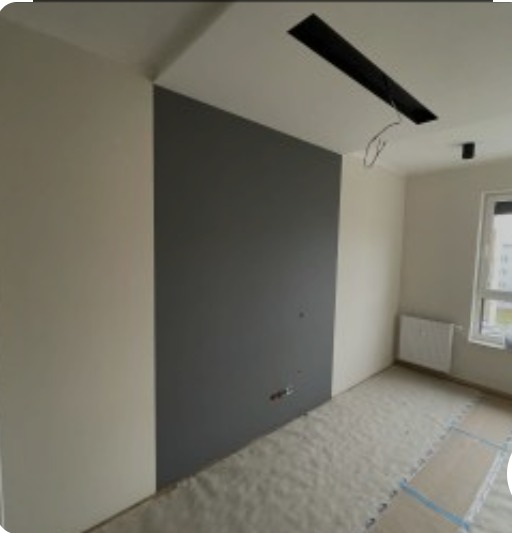 Szare wykończenie ściany w pokoju z przygotowanym podłożem pod podłogę. Widoczne gniazdka elektryczne i otwór w suficie na oświetlenie.