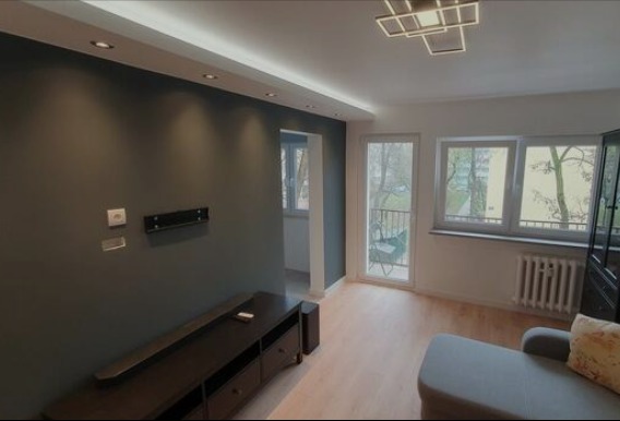 Nowoczesny salon z szarą ścianą, drewnianą szafką RTV i wyjściem na balkon. Widoczne okna z widokiem na zieleń i designerska lampa sufitowa.