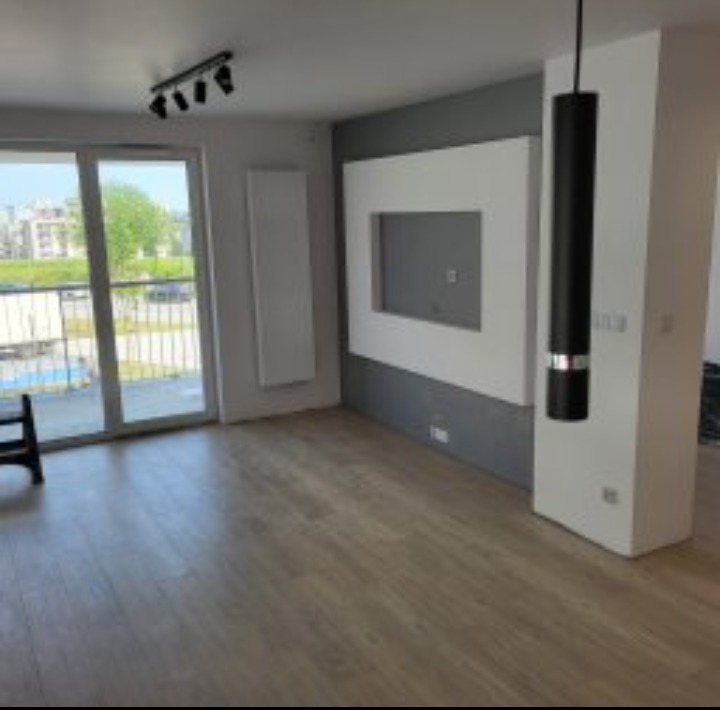 Jasne wnętrze po remoncie: parkiet, szara ściana z białą wnęką na TV, balkon z widokiem na zieleń. Nowoczesne oświetlenie sufitowe i wisząca lampa.