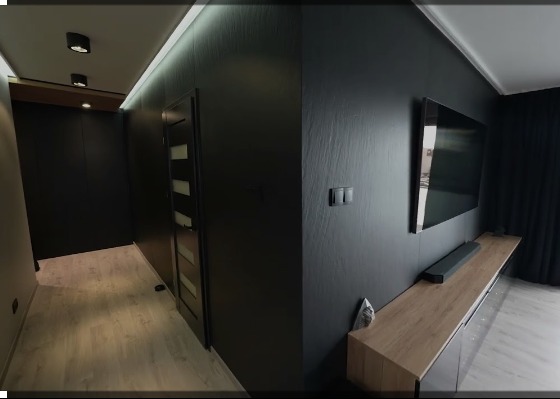 Nowoczesne wnętrze: czarne ściany z fakturą, drewnopodobna podłoga, minimalistyczna szafka RTV i telewizor na ścianie. Widoczny fragment korytarza z drzwiami.