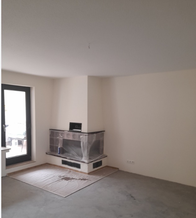 Wnętrze pokoju z kominkiem zabezpieczonym folią malarską, świeżo po remoncie. Widoczne gładkie ściany i wylewka na podłodze. Czarna rama okienna po lewej.