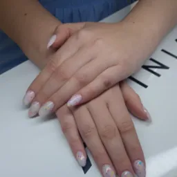 Dłonie z owalnymi paznokciami w jasnym odcieniu, zdobionymi kolorowymi drobinkami, na białym tle. Widoczny fragment niebieskiej odzieży.