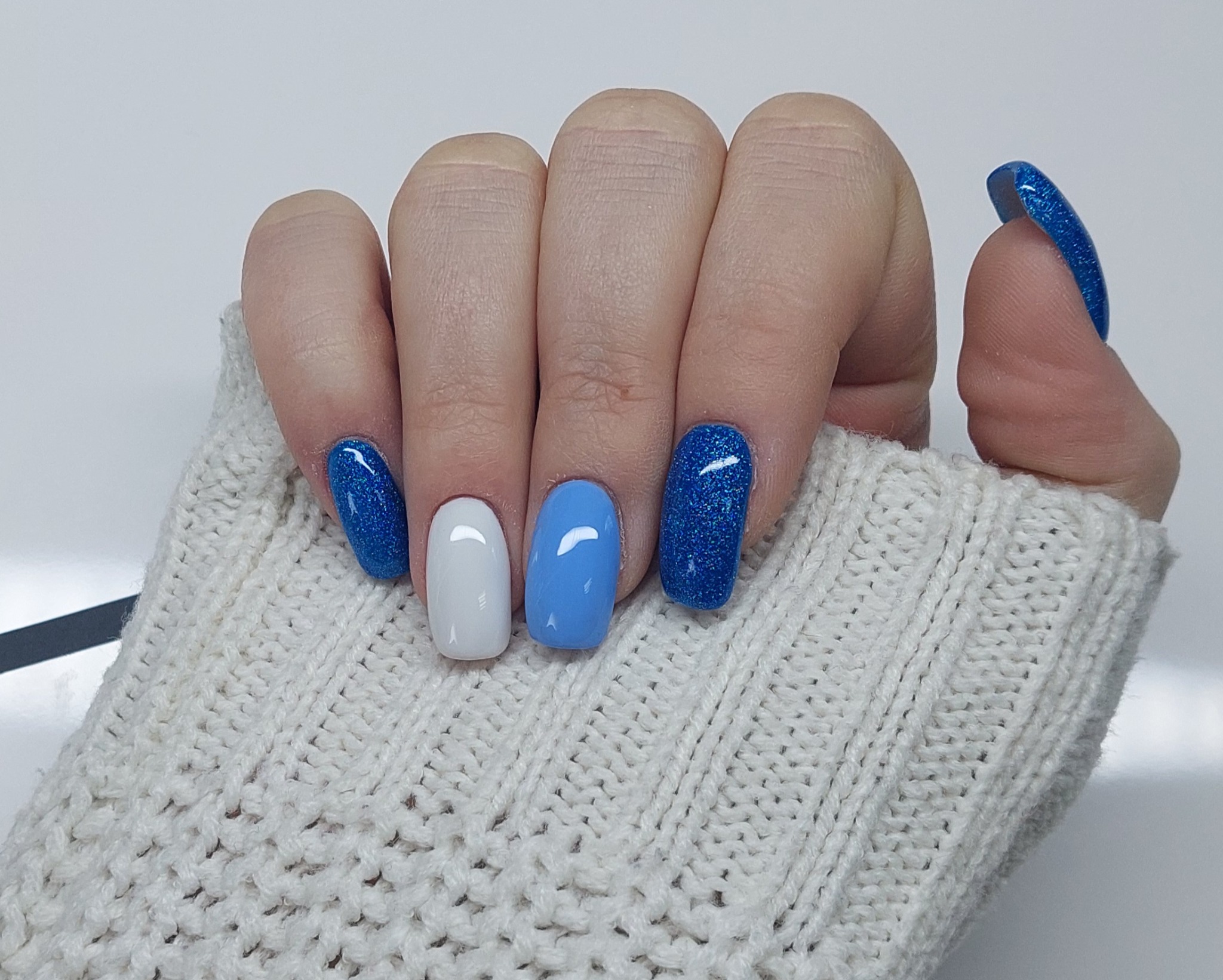 Dłoń w kremowym swetrze prezentuje paznokcie pomalowane na biało, błękitny i ciemnoniebieski brokat, z naciskiem na kontrast kolorów i faktur.