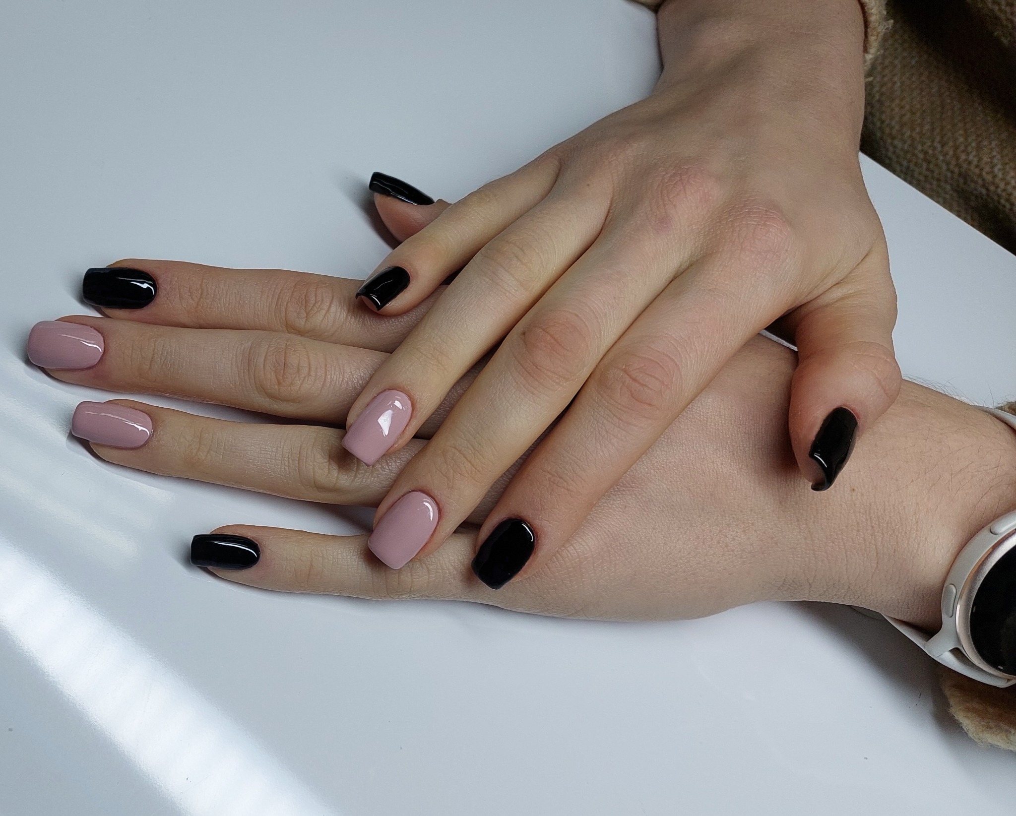 Dłonie z naprzemiennie pomalowanymi paznokciami w kolorach: pudrowy róż i czarny, na białym tle, widoczny zegarek na nadgarstku.