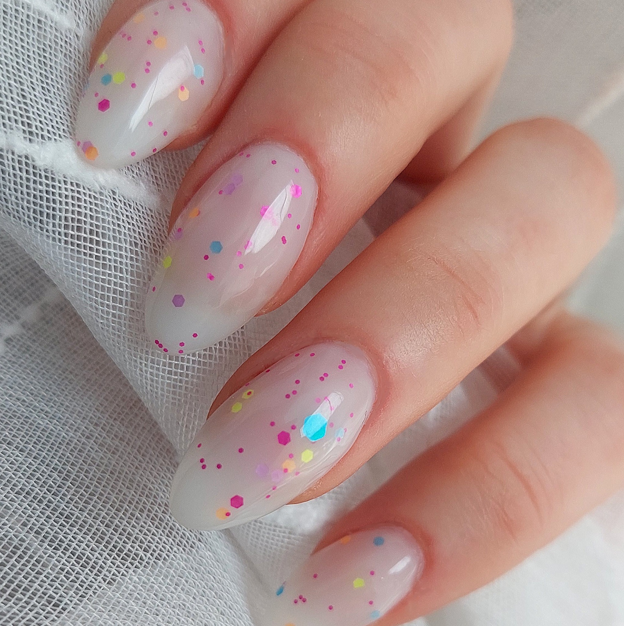 Dłoń z owalnymi paznokciami w kolorze mlecznej bieli, ozdobionymi wielokolorowymi, drobnymi kropkami i heksagonami, na tle delikatnej, białej tkaniny.