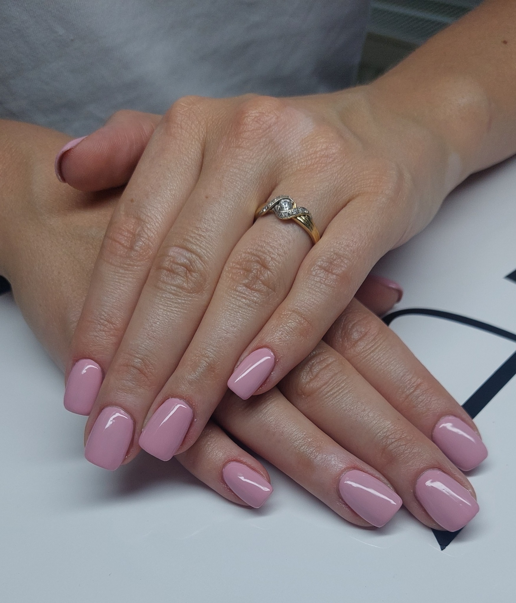 Dłonie kobiety ze świeżo pomalowanymi, kwadratowymi paznokciami w odcieniu pudrowego różu, ozdobione złotym pierścionkiem z cyrkoniami.