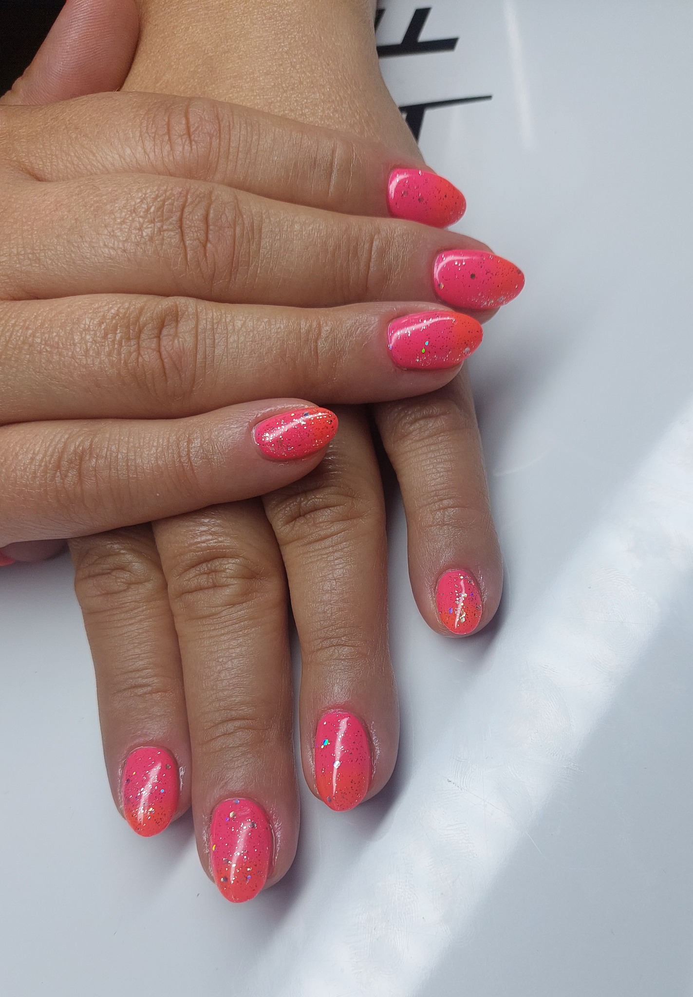 Dłonie z pomalowanymi na różowo-pomarańczowo paznokciami z brokatem, na białym tle. Manicure hybrydowy z efektem ombre i błyszczącymi drobinkami.