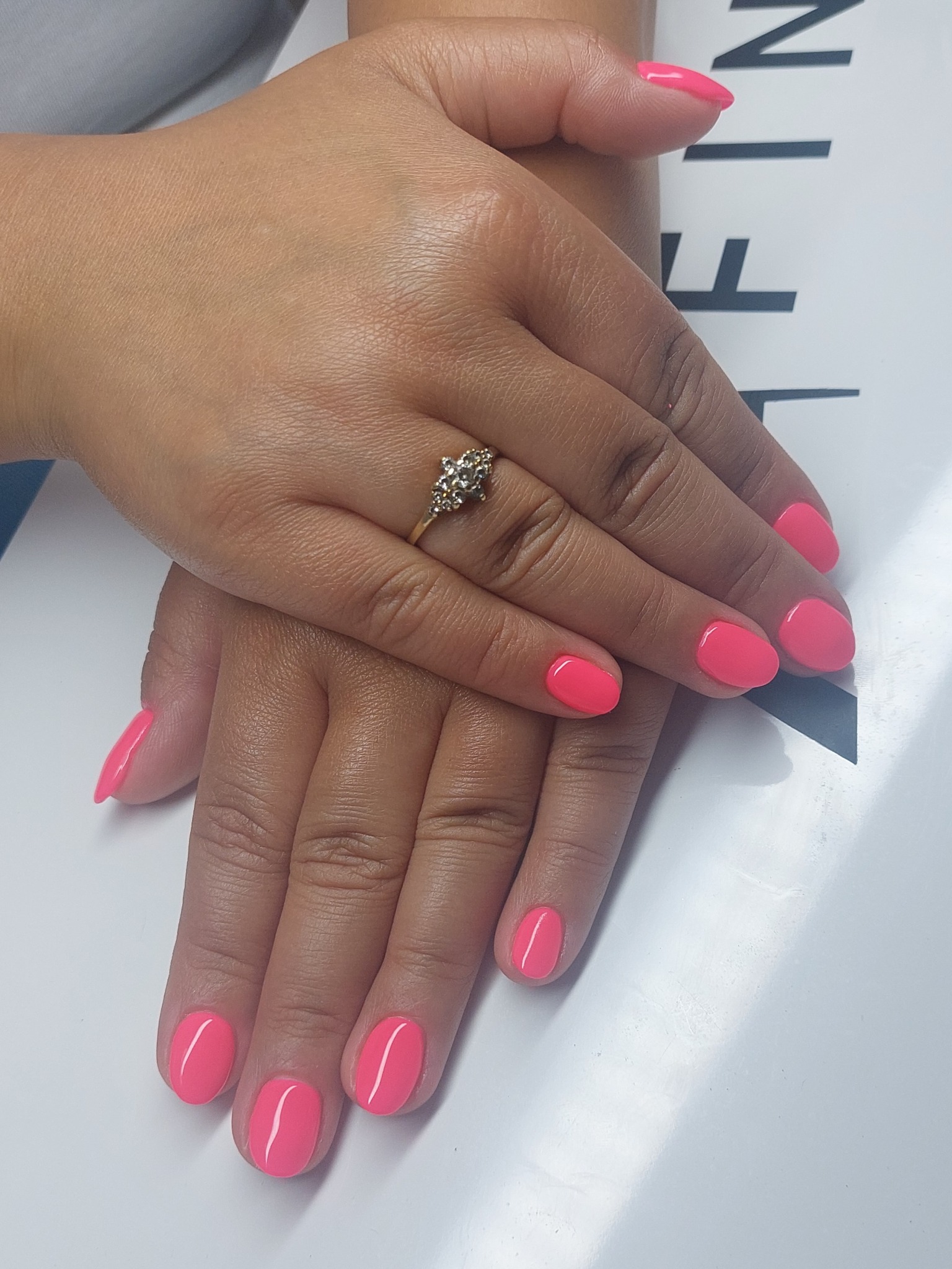 Dłonie z świeżym, neonowo-różowym manicure hybrydowym i złotym pierścionkiem na serdecznym palcu, ułożone na białym tle z czarnymi napisami.