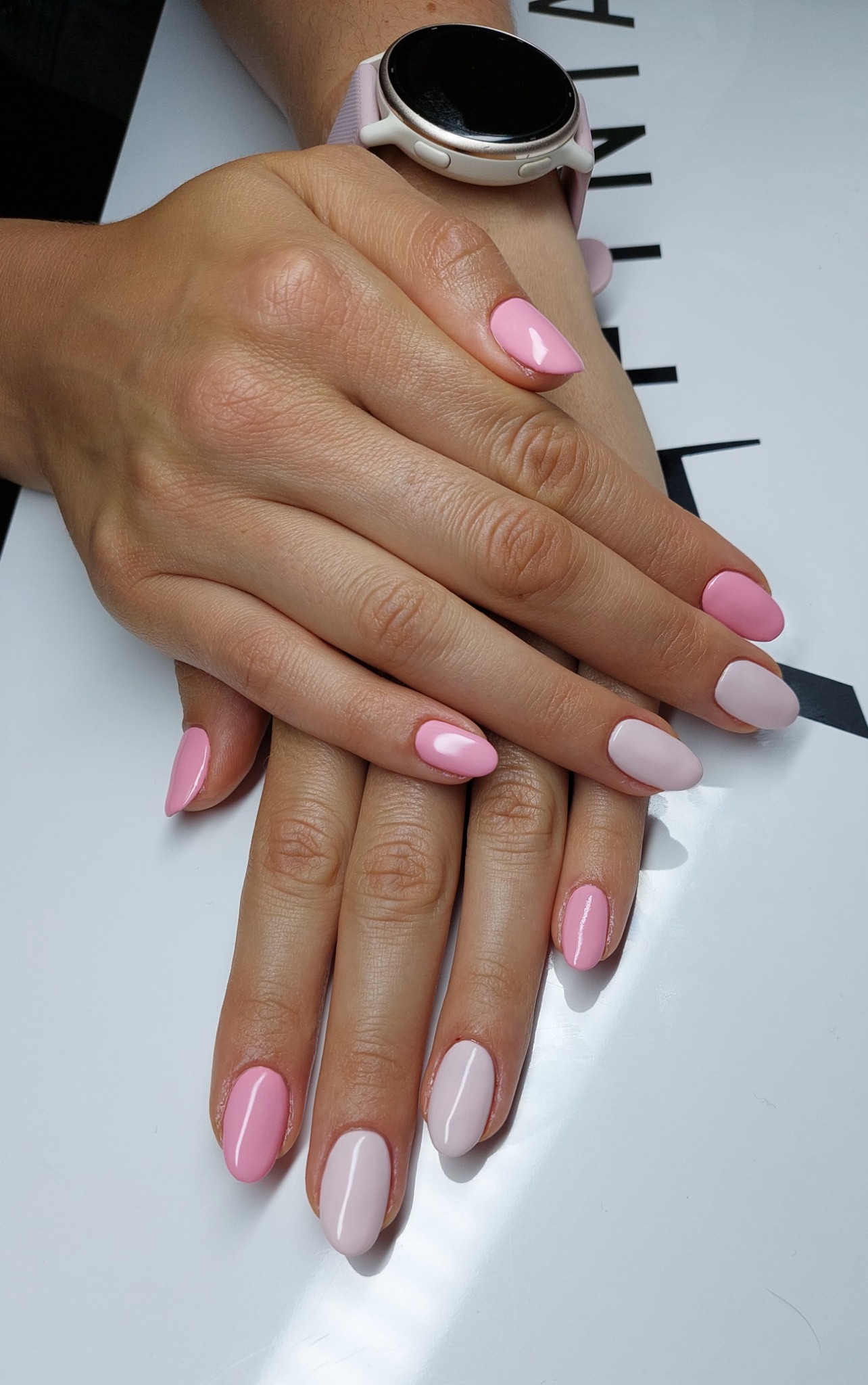 Dłonie z delikatnym, dwukolorowym manicure w odcieniach różu, na jasnym tle. Na nadgarstku widoczny smartwatch z jasną bransoletą.