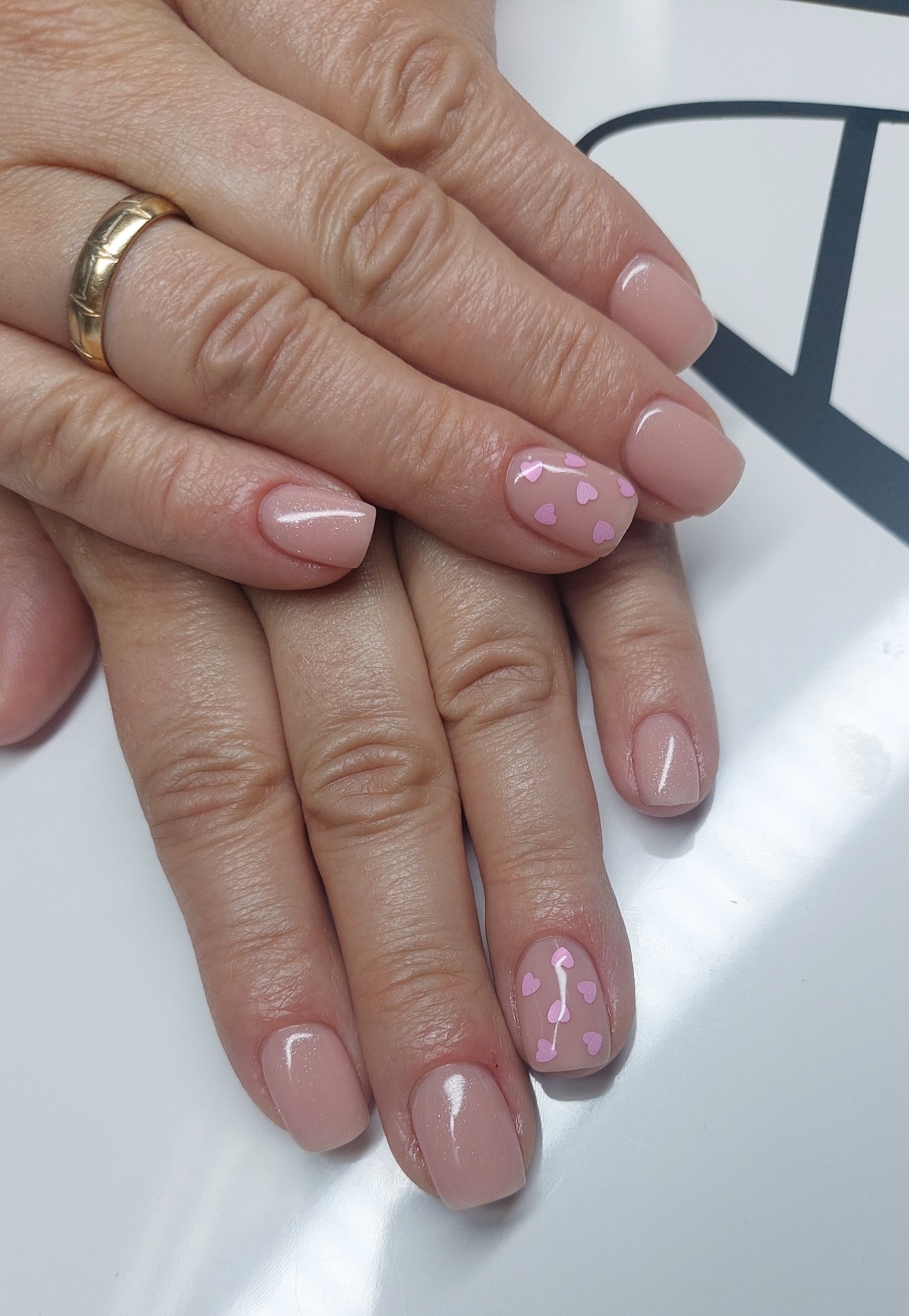 Delikatny manicure w odcieniach nude z drobnymi, różowymi serduszkami na jednym paznokciu, dłonie spoczywają na białym tle, widoczny złoty pierścionek.