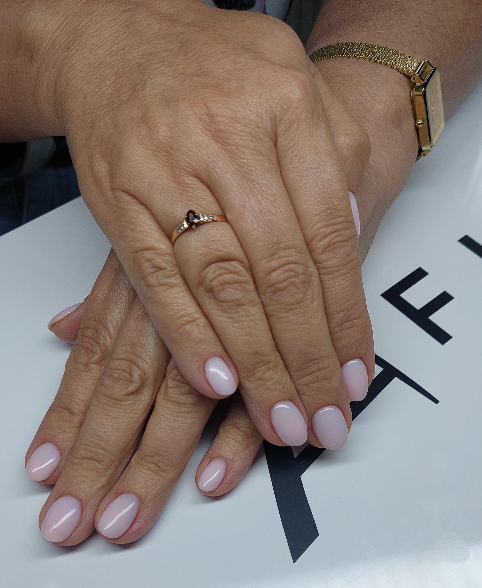 Delikatny, pudrowy manicure na dłoni z pierścionkiem i złotym zegarkiem. Krótkie, owalne paznokcie na białym tle z czarnym detalem.