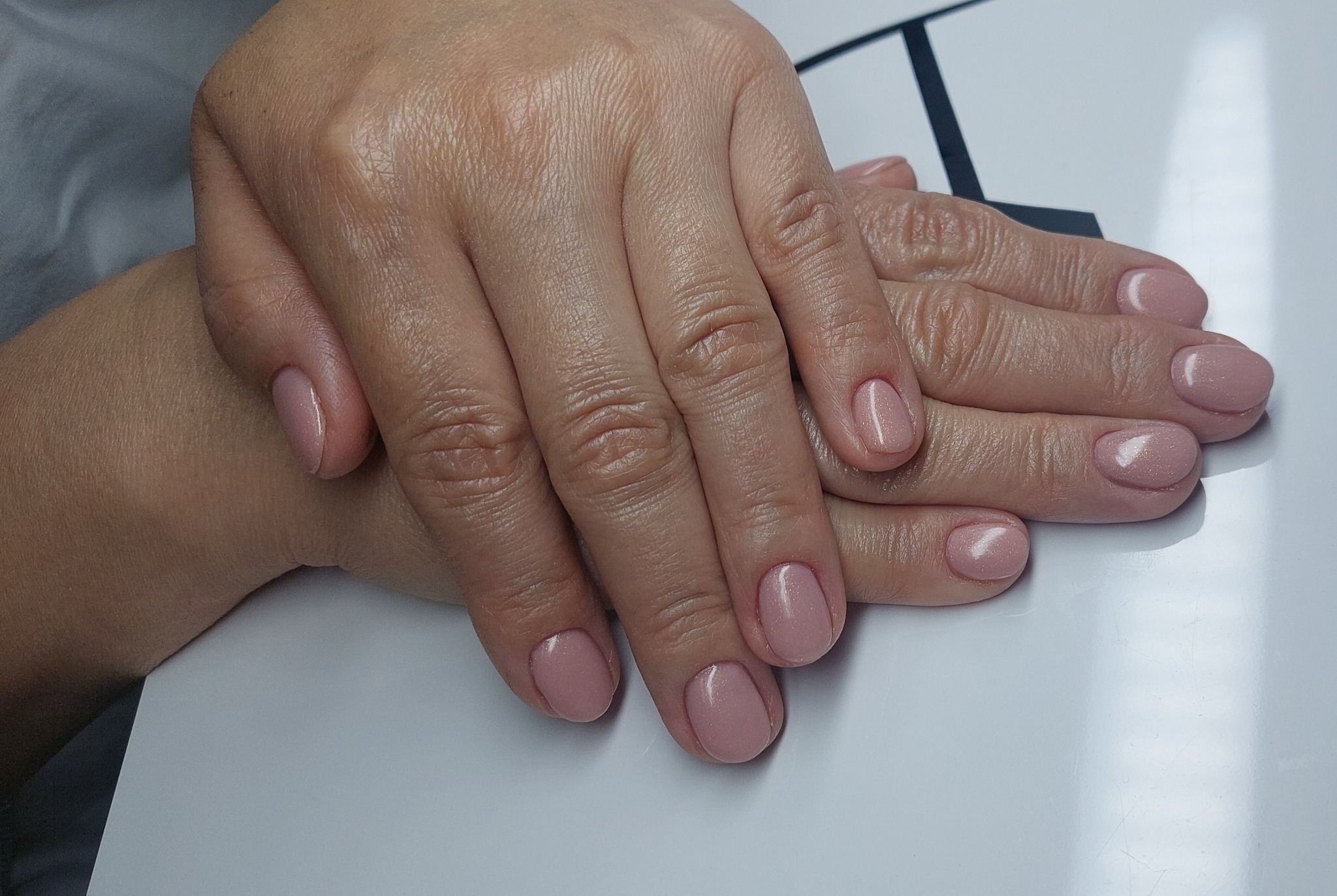 Dłonie z delikatnym, różowym manicure hybrydowym na krótkich paznokciach, ułożone na białym tle, z widoczną strukturą skóry i subtelnym połyskiem lakieru.