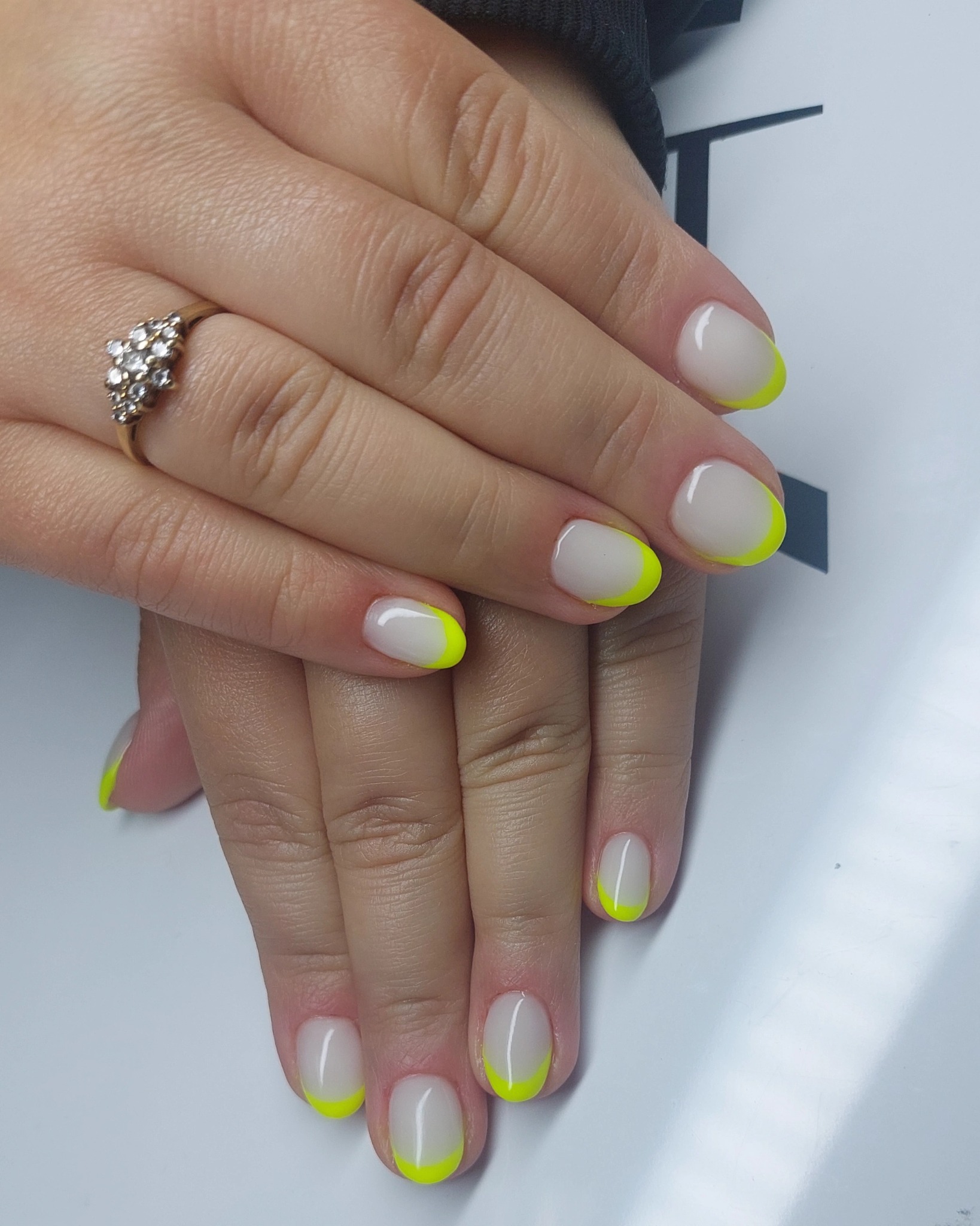 Dłonie z białym manicure z neonowo-żółtymi końcówkami, na palcu serdecznym złoty pierścionek z kamieniami, na jasnym tle.
