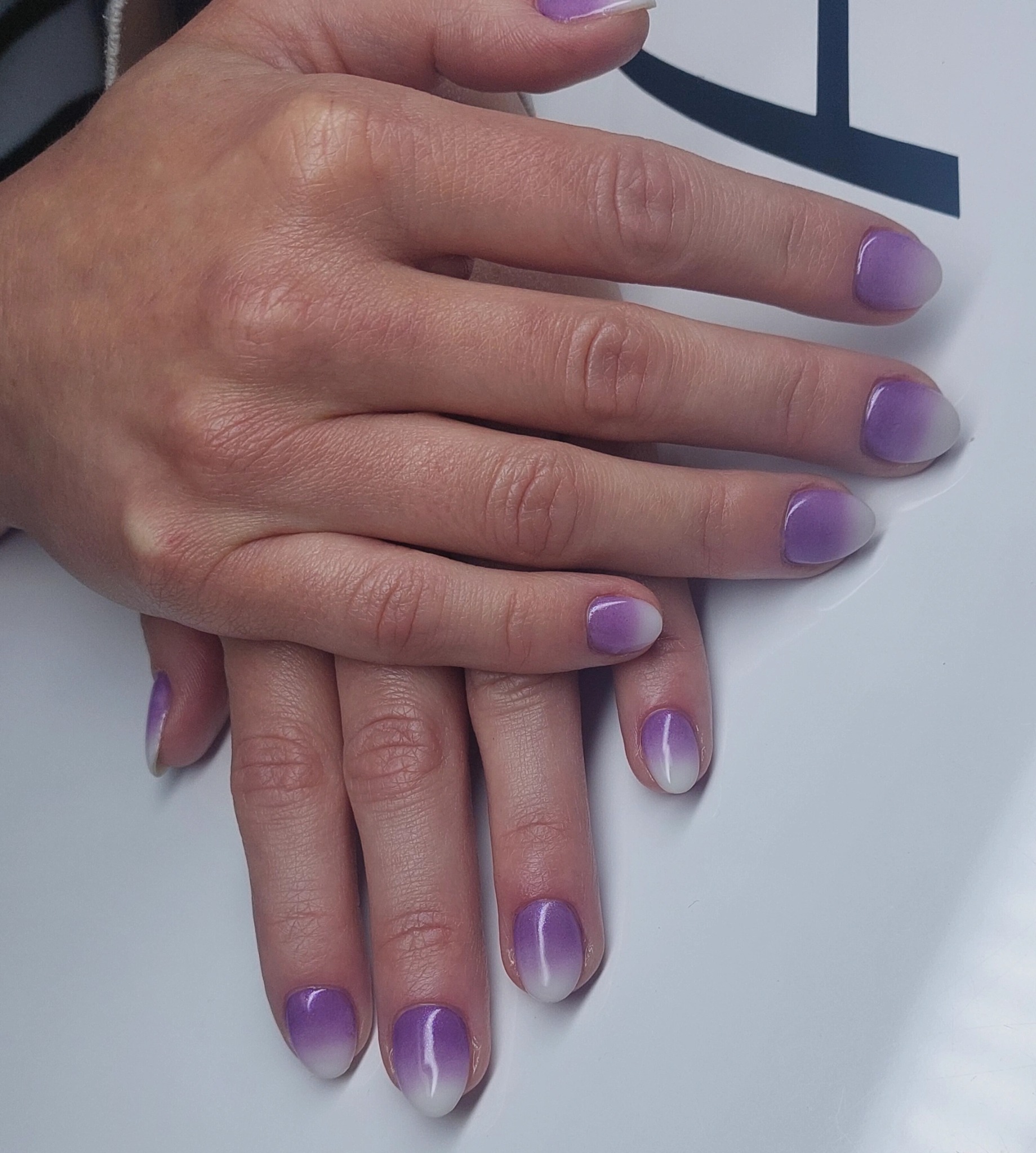 Dłonie z fioletowo-białym manicure ombre na krótkich, owalnych paznokciach. Efektowny gradient i błyszczące wykończenie na białym tle.