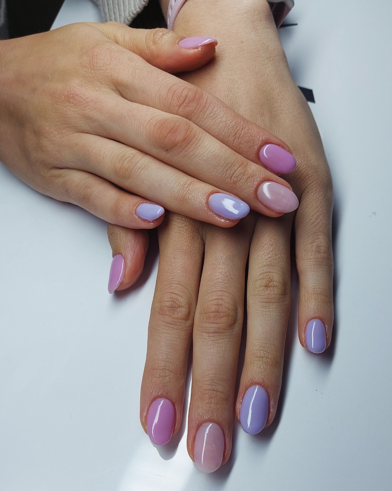 Dłonie z pastelowym manicure hybrydowym w odcieniach fioletu i różu, na białym tle. Widoczne skórki po zabiegu pielęgnacyjnym. Delikatny połysk lakieru.