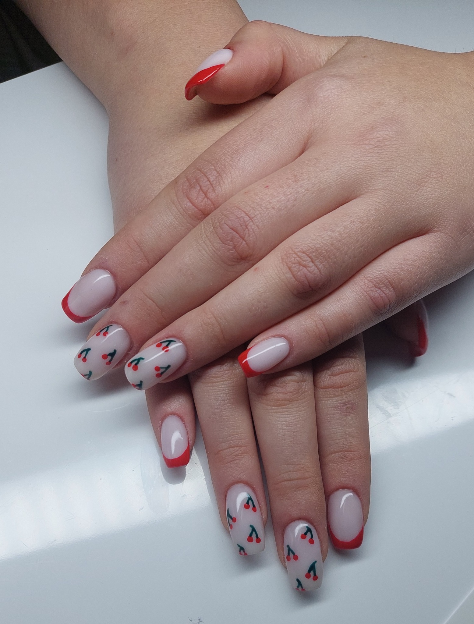 Dłonie z pomalowanymi paznokciami: french manicure z czerwoną końcówką i wzorem w wiśnie na jasnym tle. Widoczne delikatne detale skóry dłoni.