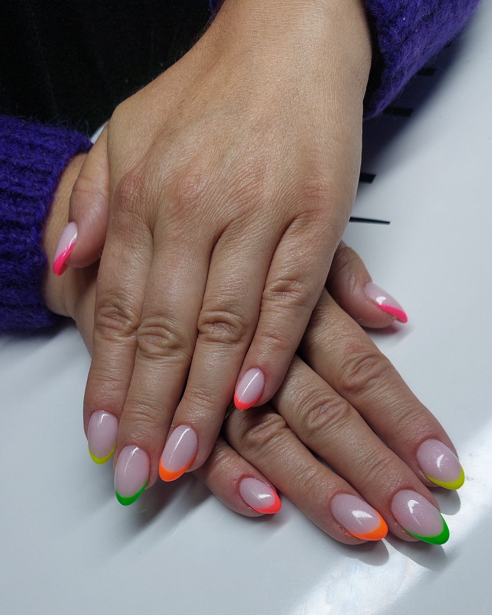 Dłonie z manicure french w neonowych kolorach: różowym, pomarańczowym, zielonym i żółtym. Detal: fioletowy sweter w tle, podkreślający kontrast barw.