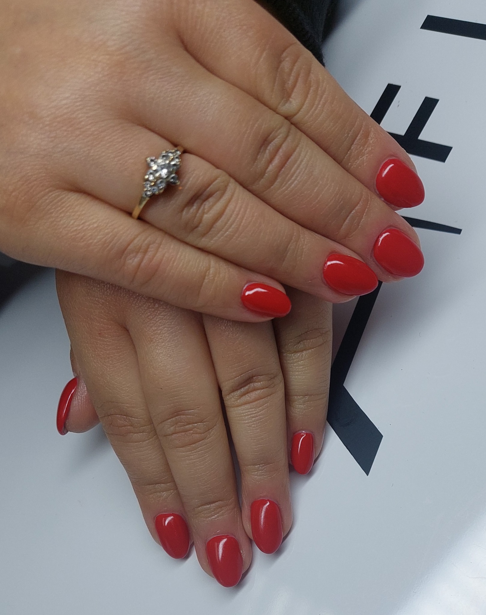 Dłonie z czerwonym manicure hybrydowym i pierścionkiem z brylantami na białym tle z czarnymi elementami graficznymi.