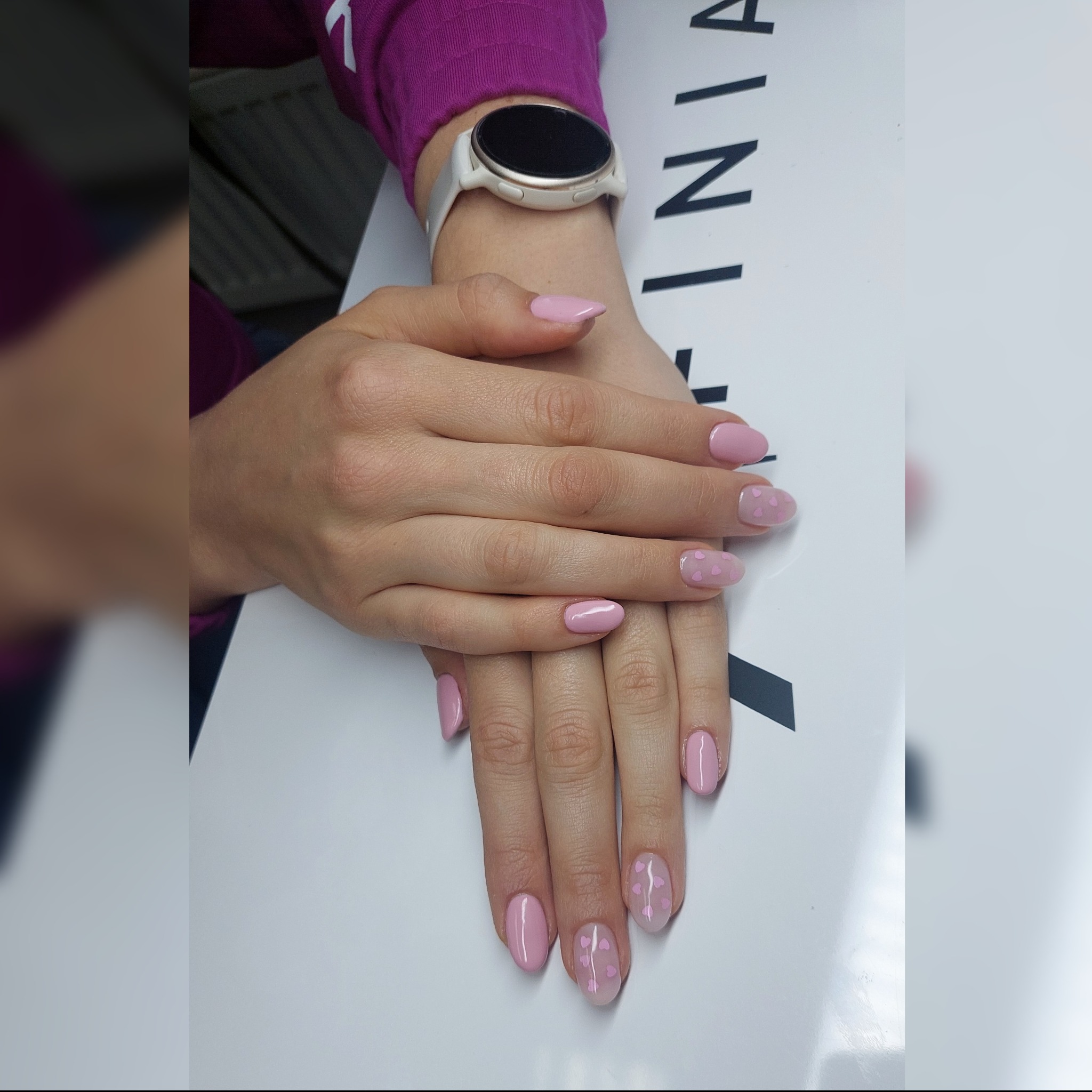 Ręce z delikatnym, różowym manicure z motywem małych serduszek na kilku paznokciach, na jasnym tle. Widoczny zegarek na nadgarstku.