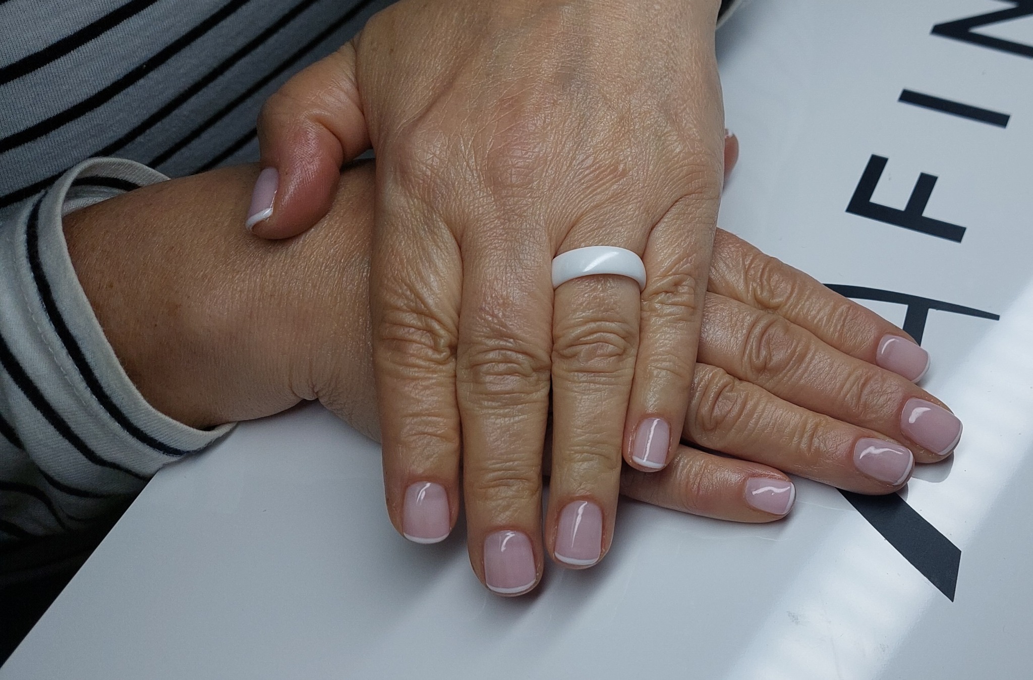 Dłonie z delikatnym manicure francuskim, biały pierścionek na palcu wskazującym, bluzka w czarno-białe paski w tle. Subtelny i elegancki wygląd.