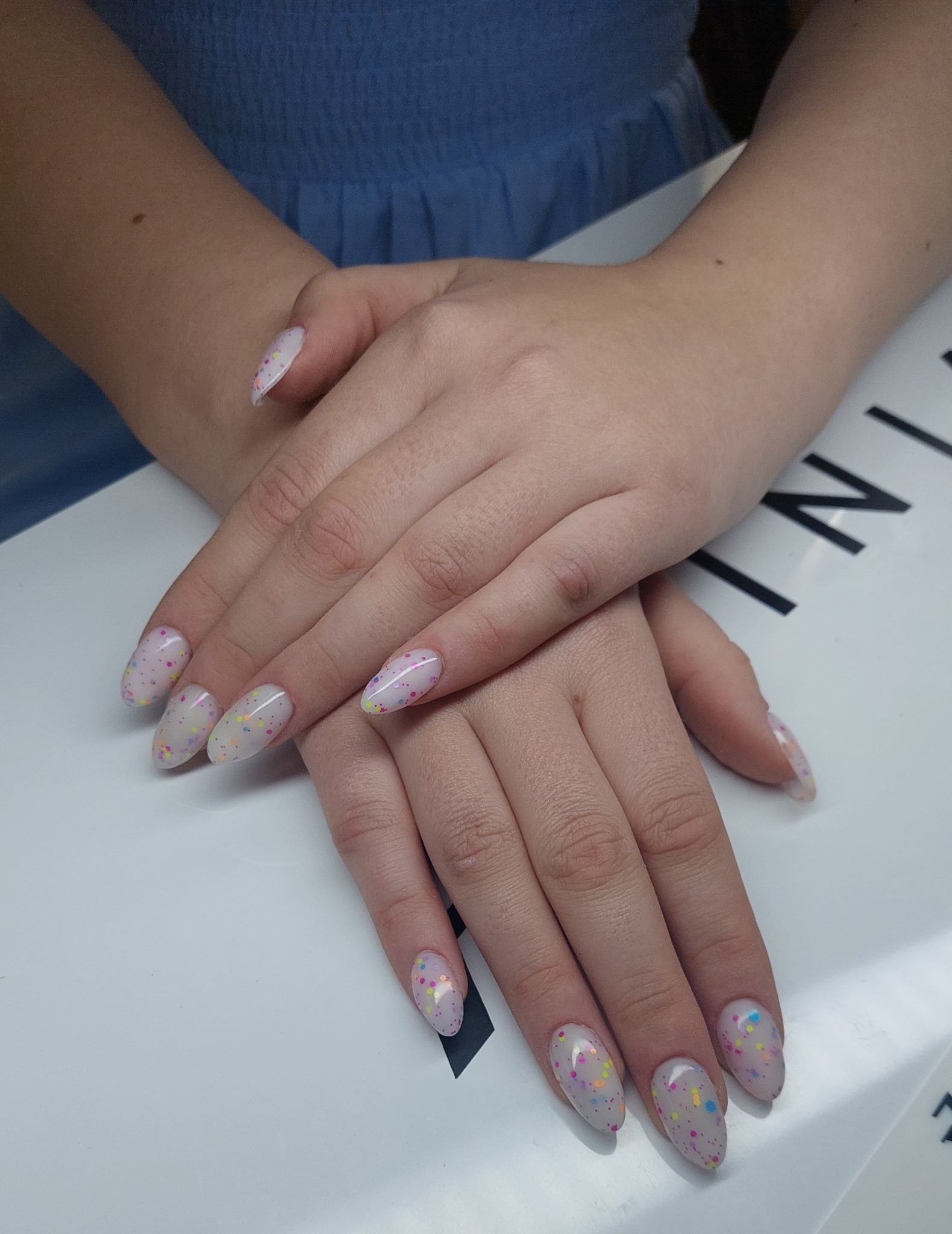 Dłonie z owalnymi paznokciami w jasnym odcieniu, zdobionymi kolorowymi drobinkami, na białym tle. Widoczny fragment niebieskiej odzieży.