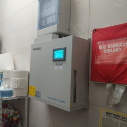 Przedsiębiorstwo Pomiarów i Automatyzacji Advanced Solutions inż. Adam Gawlik - Szary generator statyczny Var marki Sinexcel zamontowany na ścianie z kafelków, obok czerwony koc gaśniczy szklany i instrukcja BHP w kotłowniach gazowych.