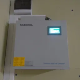 Zamontowany na ścianie szary generator mocy biernej Sinexcel z wyświetlaczem LCD i żółtymi naklejkami ostrzegawczymi.