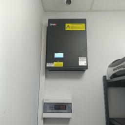 Przedsiębiorstwo Pomiarów i Automatyzacji Advanced Solutions inż. Adam Gawlik - Zamontowana szara skrzynka elektryczna ASTec z żółtą naklejką ostrzegawczą, biała tablica rozdzielcza i kamera przemysłowa na suficie w jasnym pomieszczeniu.