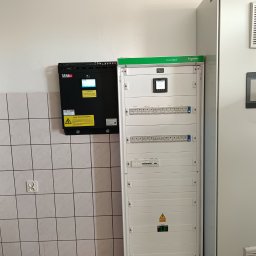 Przedsiębiorstwo Pomiarów i Automatyzacji Advanced Solutions inż. Adam Gawlik - Rozdzielnica elektryczna PrismaSeT Schneider i urządzenie ASTec na ścianie z białymi płytkami. Widoczny panel sterowania i ostrzeżenia o wysokim napięciu.