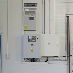 Przedsiębiorstwo Pomiarów i Automatyzacji Advanced Solutions inż. Adam Gawlik - Zamontowana szafa sterownicza SINEEXCEL z panelem kontrolnym i wyłącznikiem awaryjnym na białej ścianie z blachy trapezowej. Widoczne przewody i rury instalacyjne.