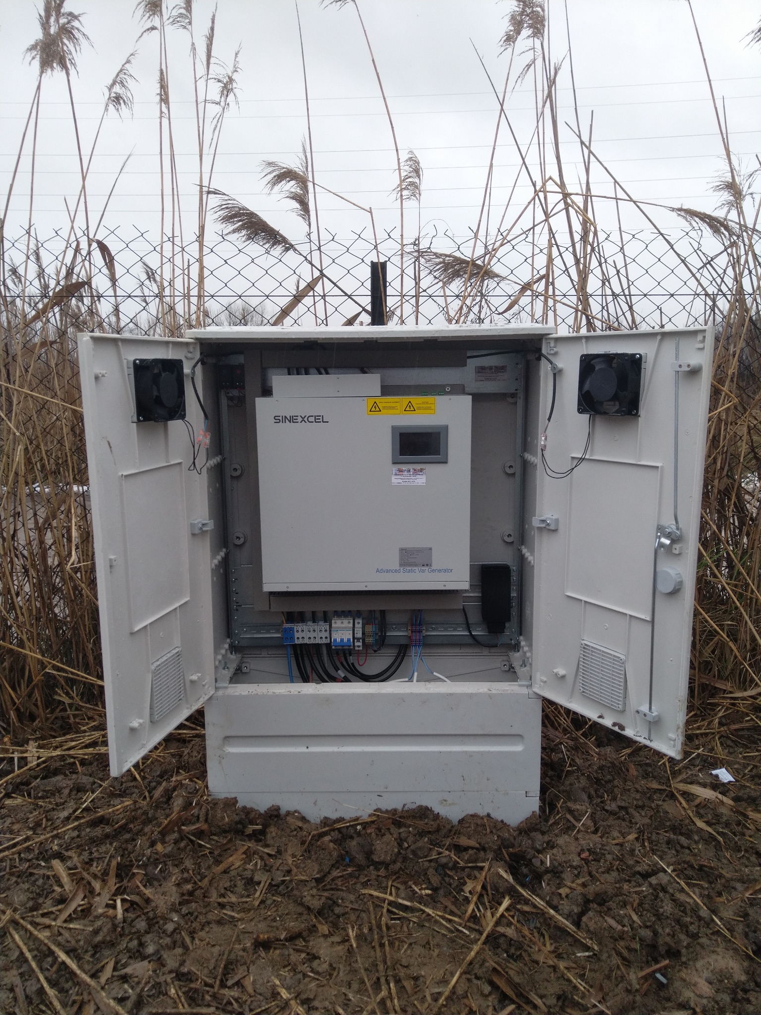 Otwarta szafa sterownicza Sinexcel Advanced Static Var Generator umieszczona na zewnątrz, w otoczeniu błotnistej ziemi i wysokich traw, z widocznymi elementami instalacji elektrycznej wewnątrz.