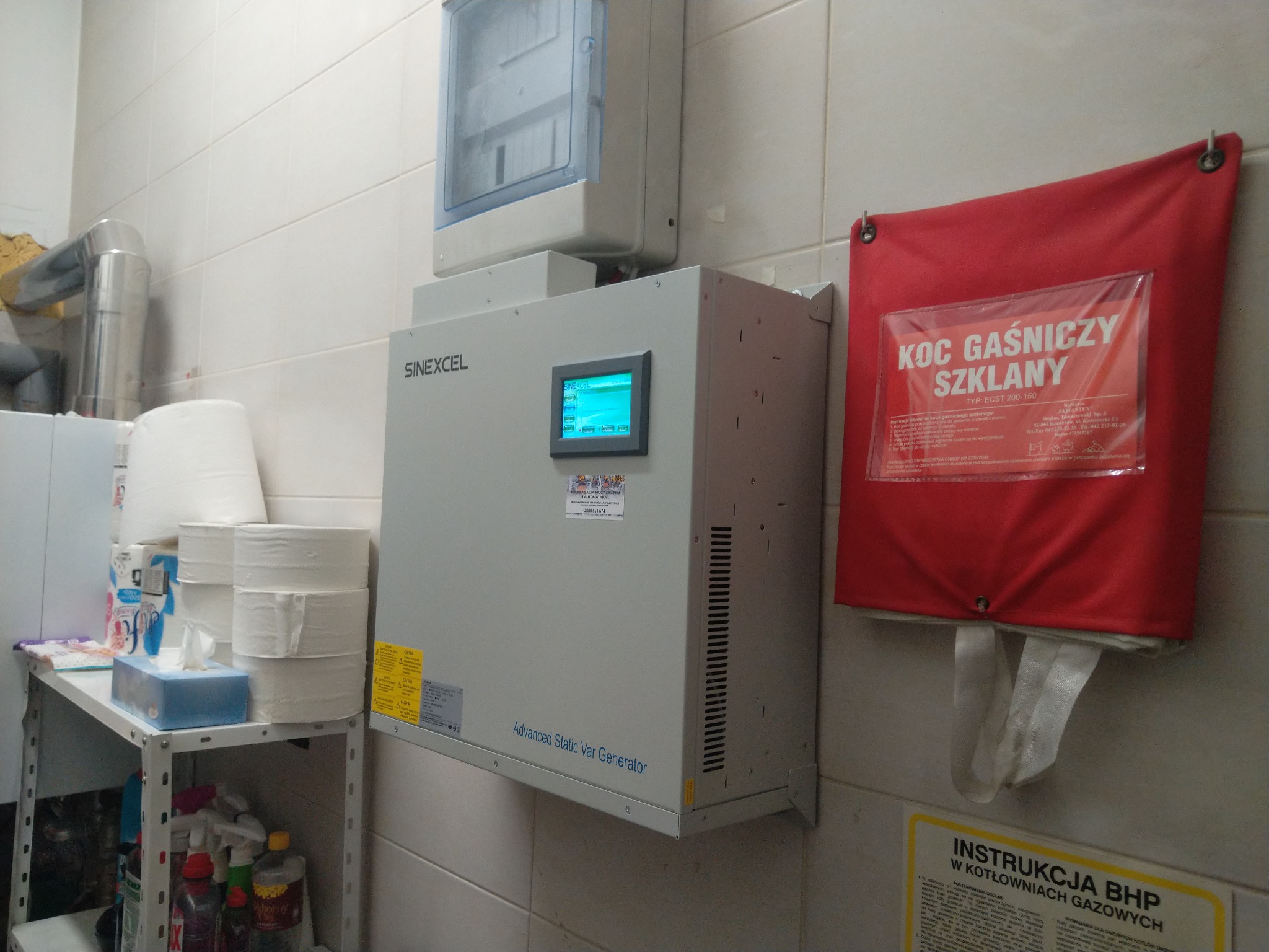 Szary generator statyczny Var marki Sinexcel zamontowany na ścianie z kafelków, obok czerwony koc gaśniczy szklany i instrukcja BHP w kotłowniach gazowych.