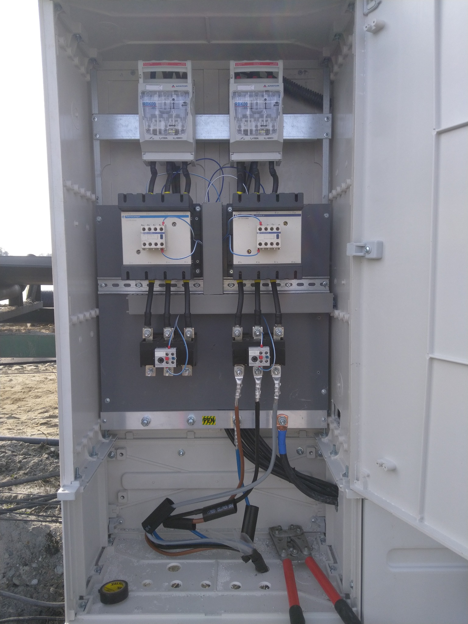 Otwarta, biała szafa elektryczna z dwoma licznikami APATOR RBK00, stycznikami LC1D115004, zabezpieczeniami i okablowaniem; na dnie szafy leżą obcęgi z czerwonymi rączkami oraz żółta miarka.