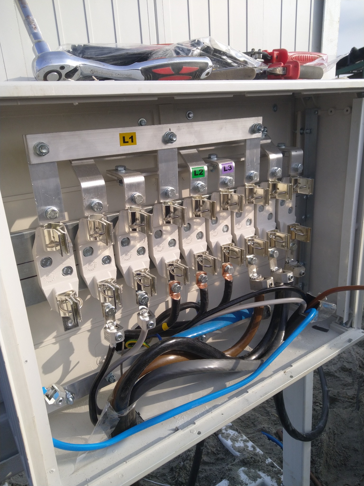 Otwarta skrzynka elektryczna z zamontowanymi szynami zbiorczymi oznaczonymi L1, L2, L3, widoczne kable i narzędzia na górze skrzynki.