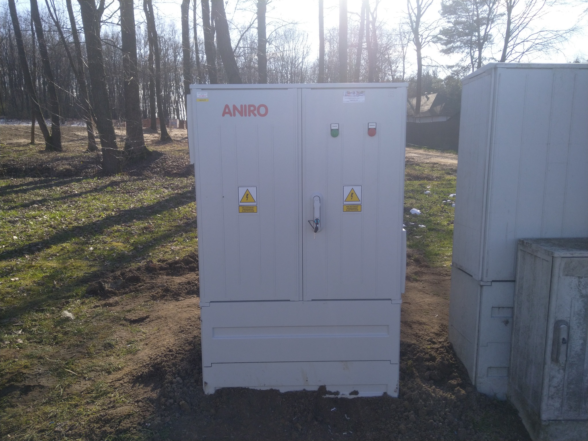 Zewnętrzna szafa elektryczna firmy ANIRO z ostrzeżeniami o napięciu, zainstalowana na trawiastym terenie w pobliżu drzew.