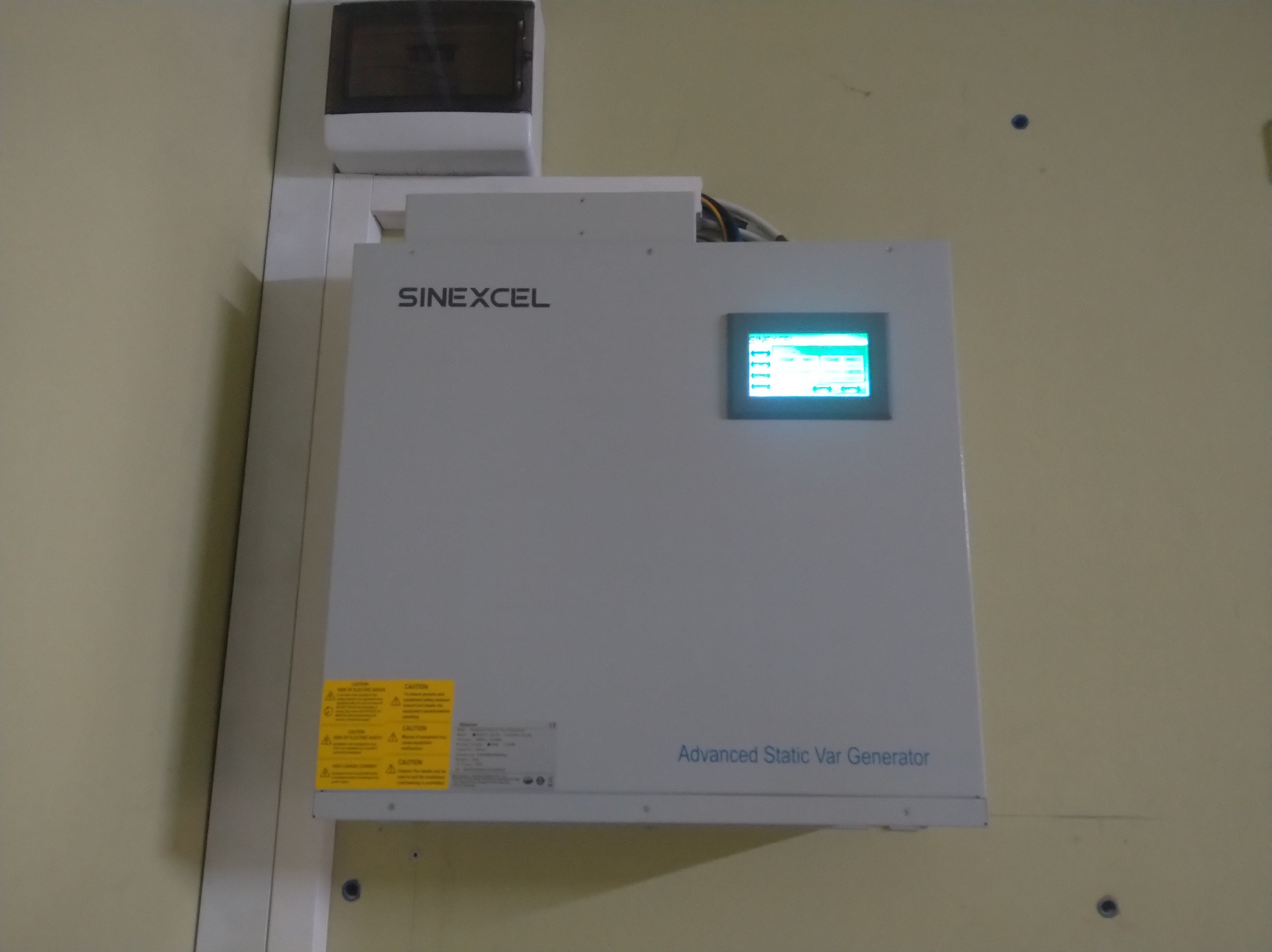 Zamontowany na ścianie szary generator mocy biernej Sinexcel z wyświetlaczem LCD i żółtymi naklejkami ostrzegawczymi.