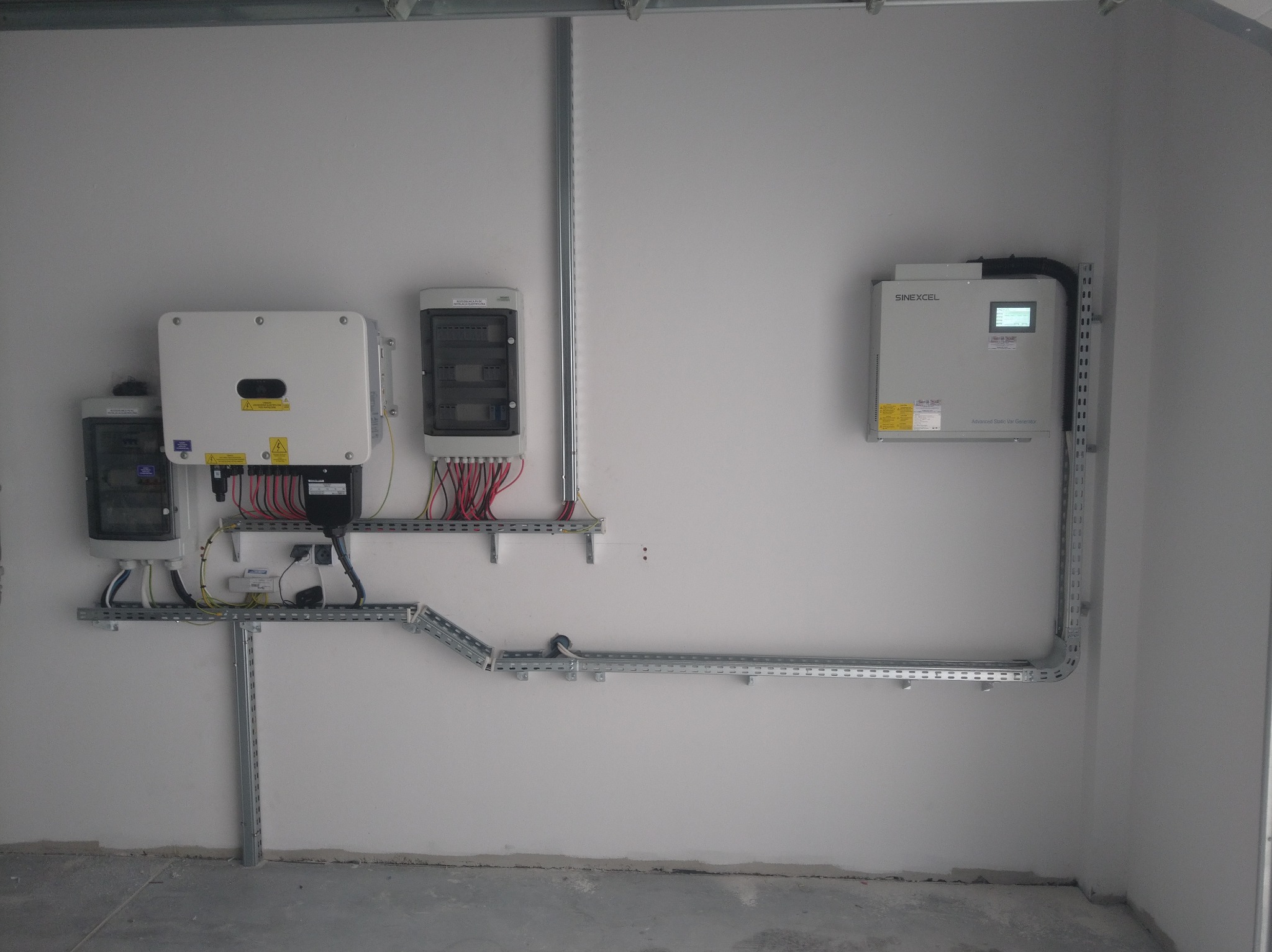 Zestawienie urządzeń elektrycznych: inwerter Sinexcel, rozdzielnice, zabezpieczenia, kable prowadzone w metalowych korytkach, zamontowane na ścianie w pomieszczeniu technicznym.