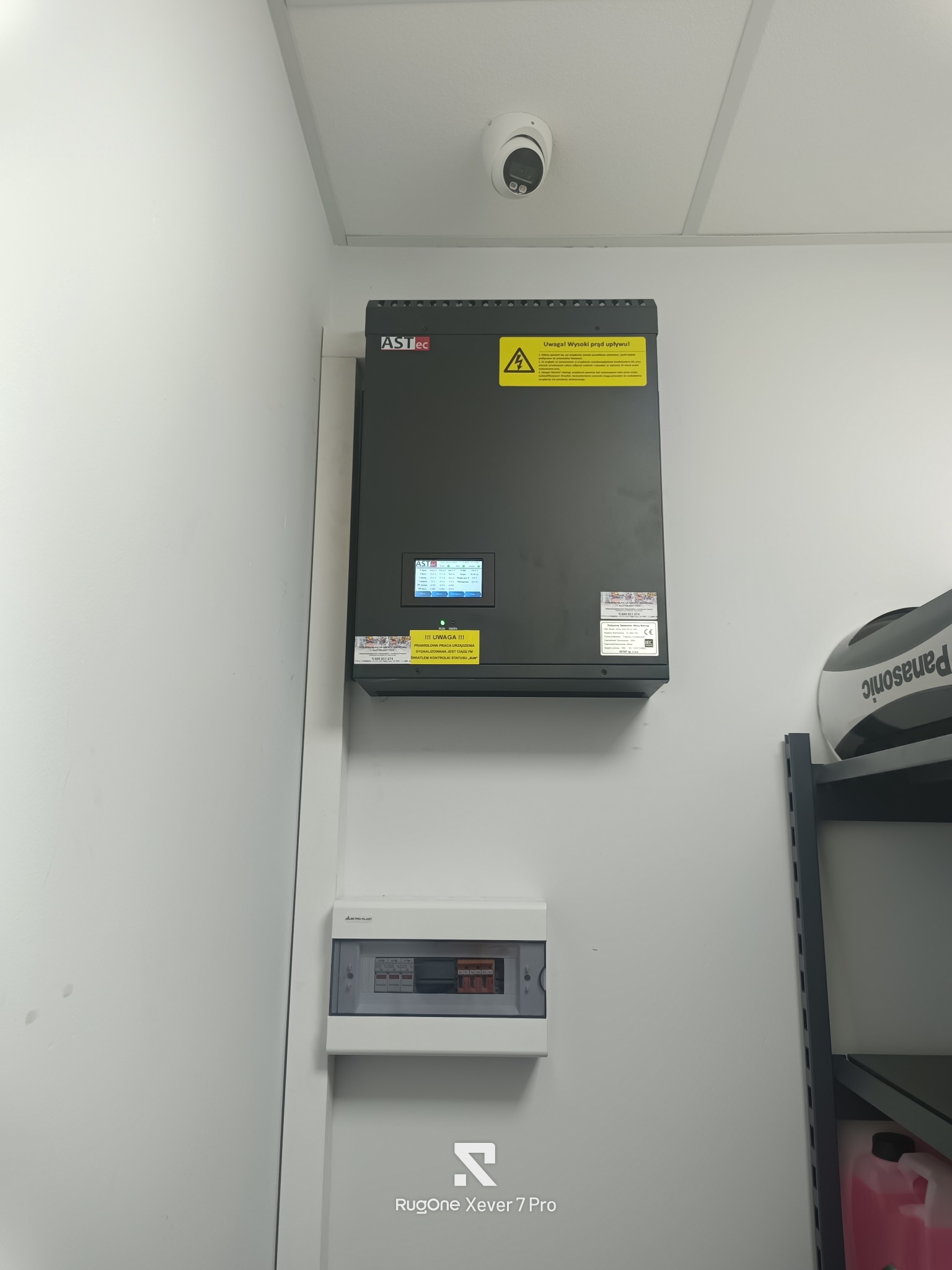 Zamontowana szara skrzynka elektryczna ASTec z żółtą naklejką ostrzegawczą, biała tablica rozdzielcza i kamera przemysłowa na suficie w jasnym pomieszczeniu.