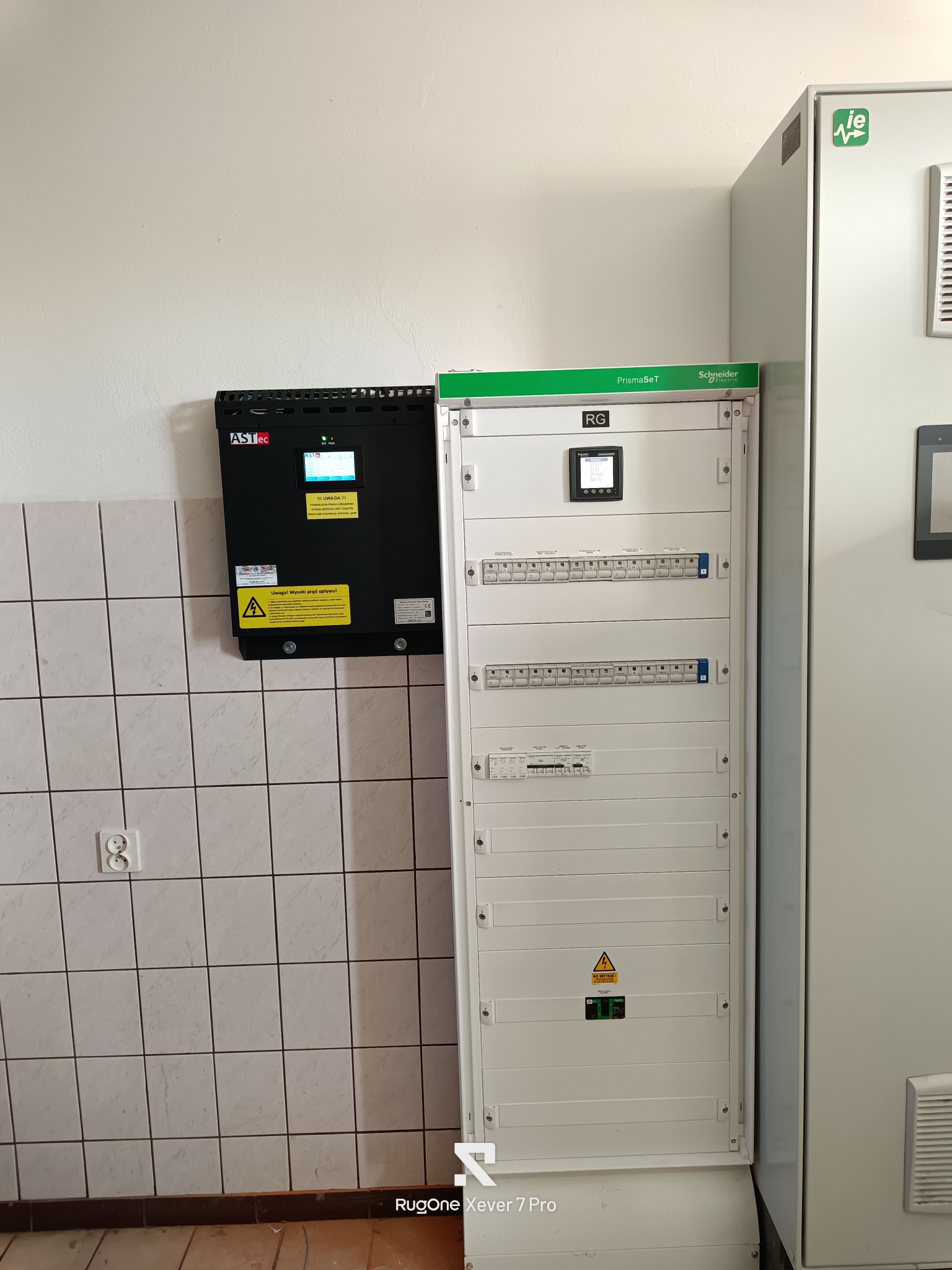 Rozdzielnica elektryczna PrismaSeT Schneider i urządzenie ASTec na ścianie z białymi płytkami. Widoczny panel sterowania i ostrzeżenia o wysokim napięciu.