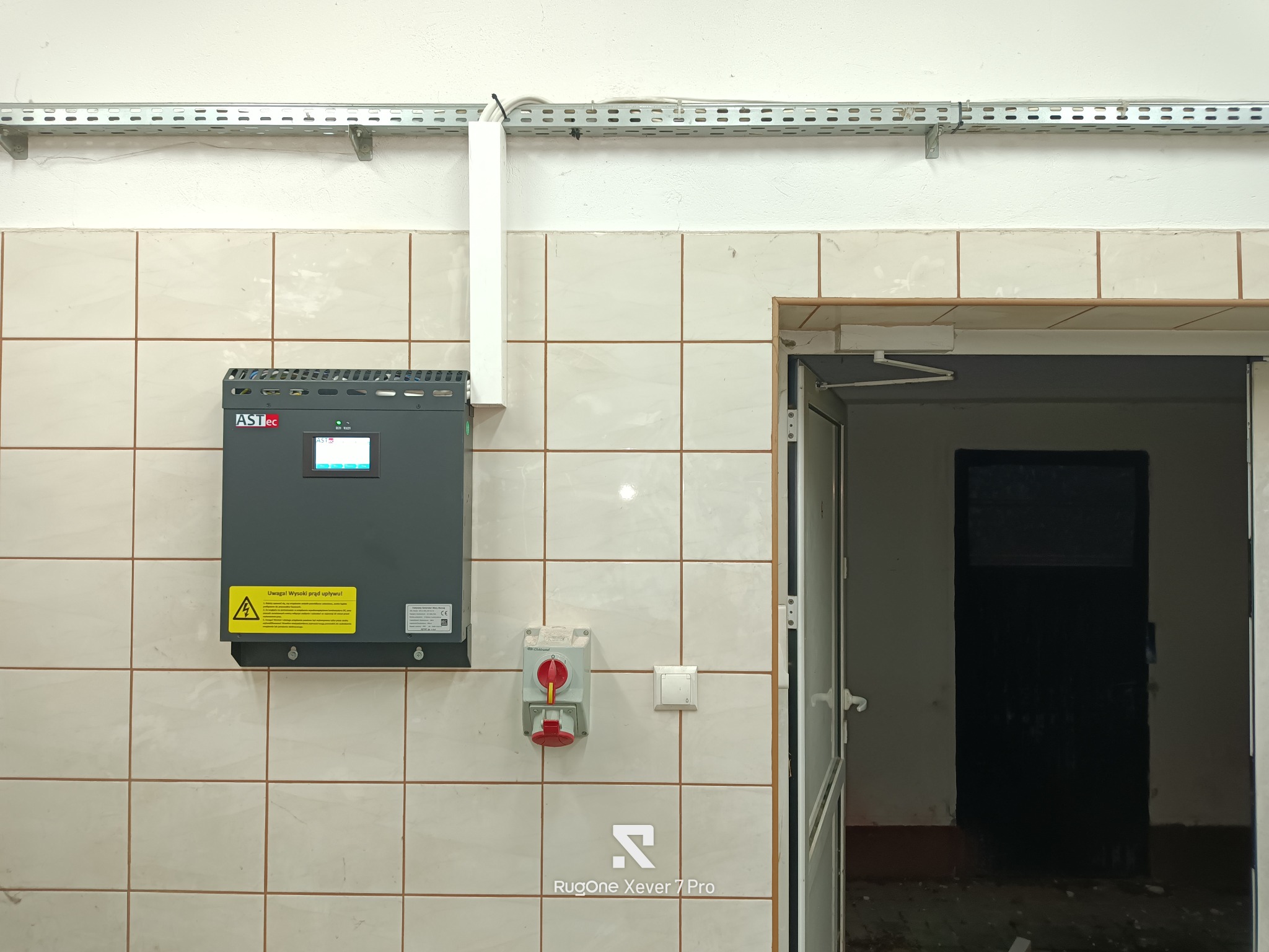 Szara skrzynka elektryczna ASTec z żółtą naklejką ostrzegawczą, zamontowana na kafelkowanej ścianie, obok wyłącznika bezpieczeństwa i gniazda. Widoczne koryto kablowe.