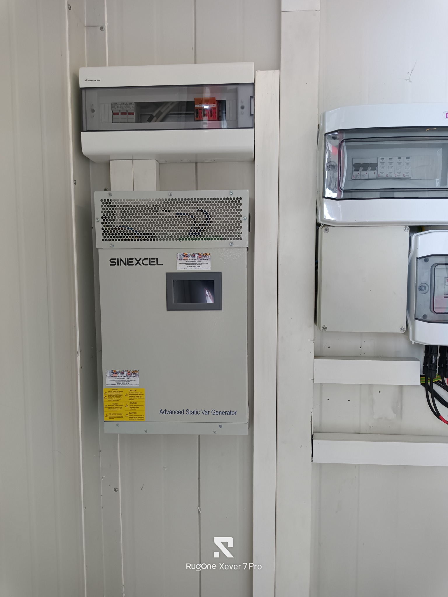 Szara szafa sterownicza Sinexcel z perforowaną górą i wyświetlaczem, zamontowana obok białych skrzynek z bezpiecznikami na ścianie z paneli. Widoczne naklejki ostrzegawcze.