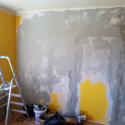 Bizoń Rafał usługi remontowo budowlane - Ściana w trakcie przygotowania do malowania, widoczne fragmenty starej żółtej farby i nałożona szara warstwa, drabina aluminiowa oparta o ścianę, obok wiadro z narzędziami.